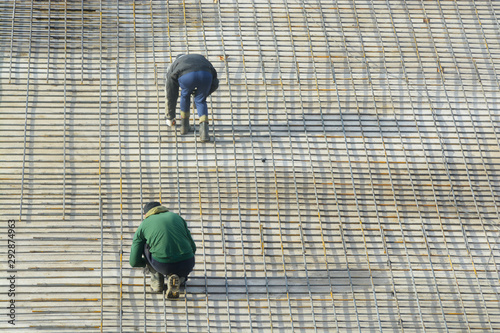Workers bind the reinforcing bars, forming a grid. In the rays of the setting sun - a warm shade of light and long shadows. This is preparation for pouring concrete Foundation of the building.