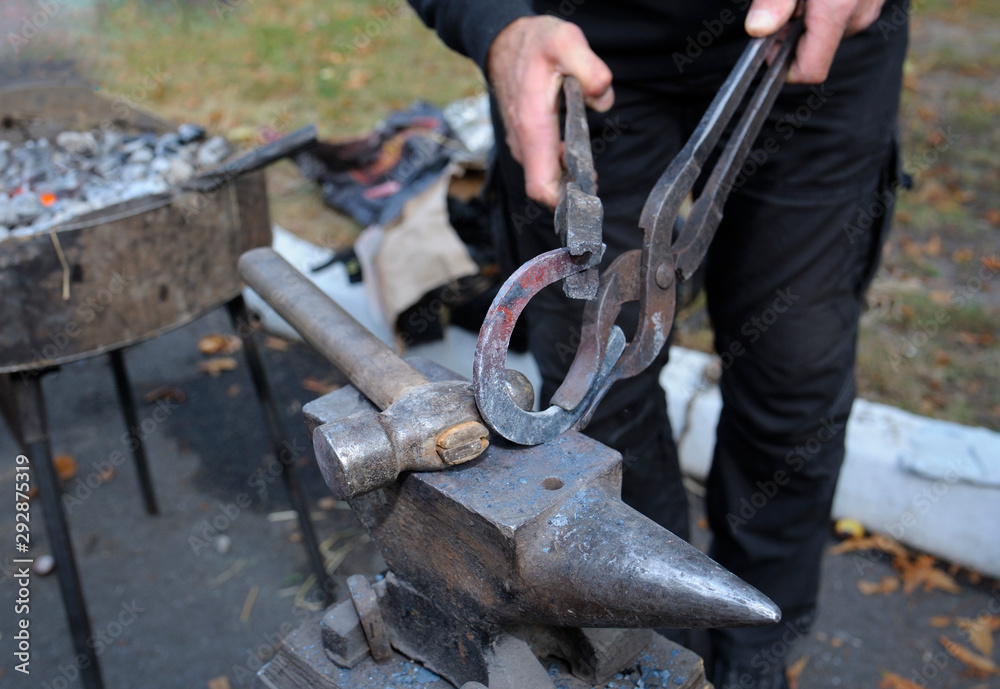At the smithy workshop. Blacksmith’s hands holding forceps and a hammer forging a metal billet, horseshoe, on an anvil At the smithy workshop. Blacksmith’s hands holding forceps and a hammer forging a metal billet, horseshoe, on an anvil