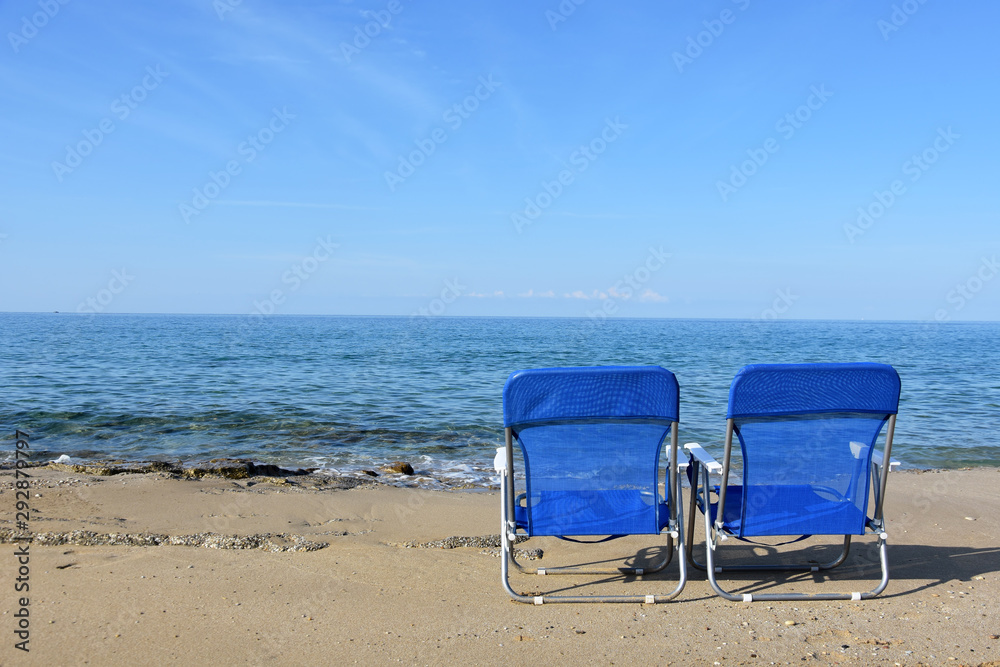 Looking at calm turquoise sea two empty chairs on sandy beach; serene sunny morning with blue sky; Ionian coast, Greece