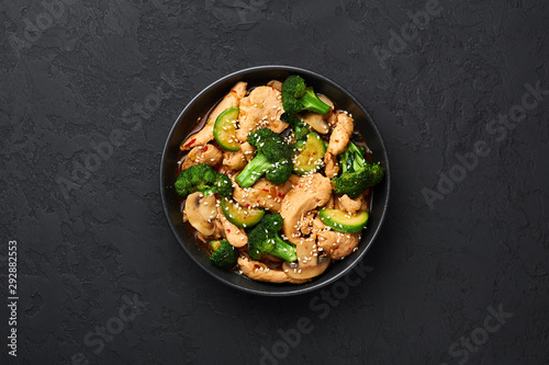 Hunan Chicken in black bowl at dark slate background. Hunan Chicken is chinese or indo-chinese cuisine takeaway dish with broccoli, zucchini, shiitake mushrooms and hunan sauce. Copy space. Top view