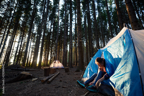 Morning camping in the forest. Young woman sitting in blue tourist tent, tying the shoe laces, getting ready to hike. On background forest, woods and white tent. Tourism active lifestyle concept