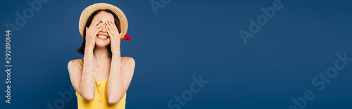 smiling girl in straw hat with hands on eyes isolated on blue
