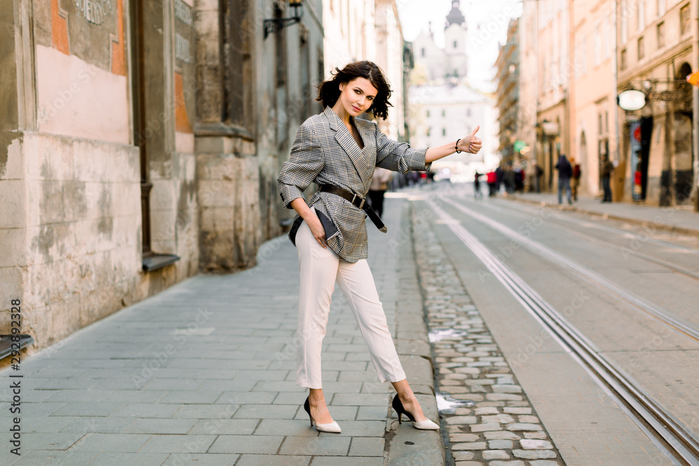 Young sexy woman in white pants and jacket hailing a taxi standing on the road, young female with hand gesture stopping taxi in urban setting, businesswoman calling taxi auto Young sexy woman in white pants and jacket hailing a taxi standing on the road, young female with hand gesture stopping taxi in urban setting, businesswoman calling taxi auto
