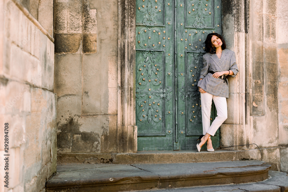 fashion shot of pretty young woman near old ancient door, she is looking at camera, wearing stylish jacket and white pants