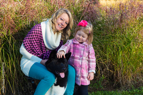 Horizontal view of smiling blond young woman sitting holding her black and white American Akita tight with her four-year old daughter petting the dog during a sunny fall morning