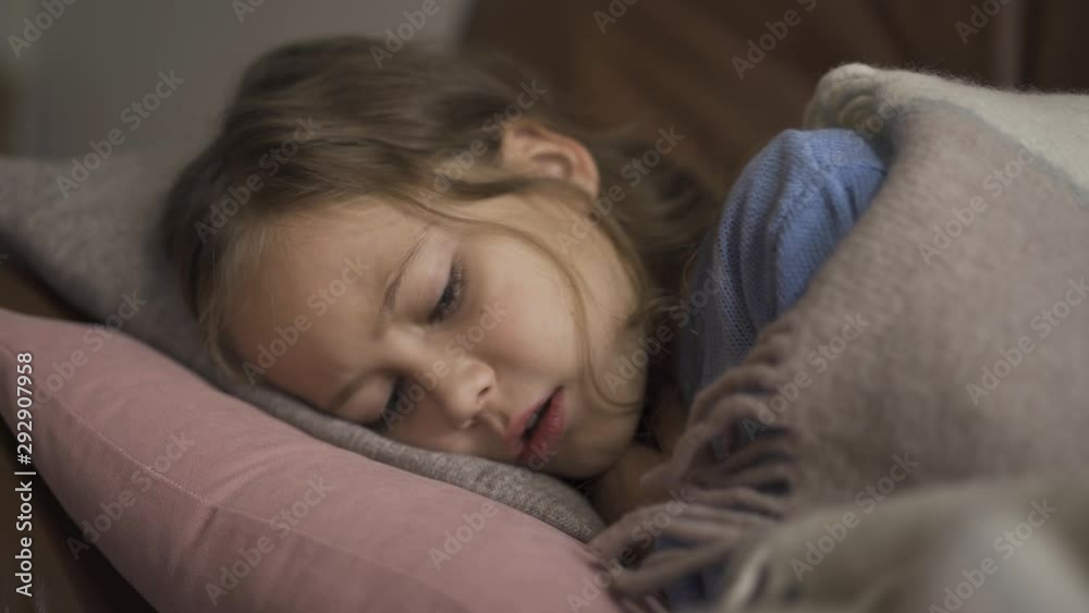 Sick young caucasian girl coughing while lying under blanket at home. The child has fever. Concept of health, illness, sickness, common cold, treatment Sick young caucasian girl coughing while lying under blanket at home. The child has fever. Concept of health, illness, sickness, common cold, treatment