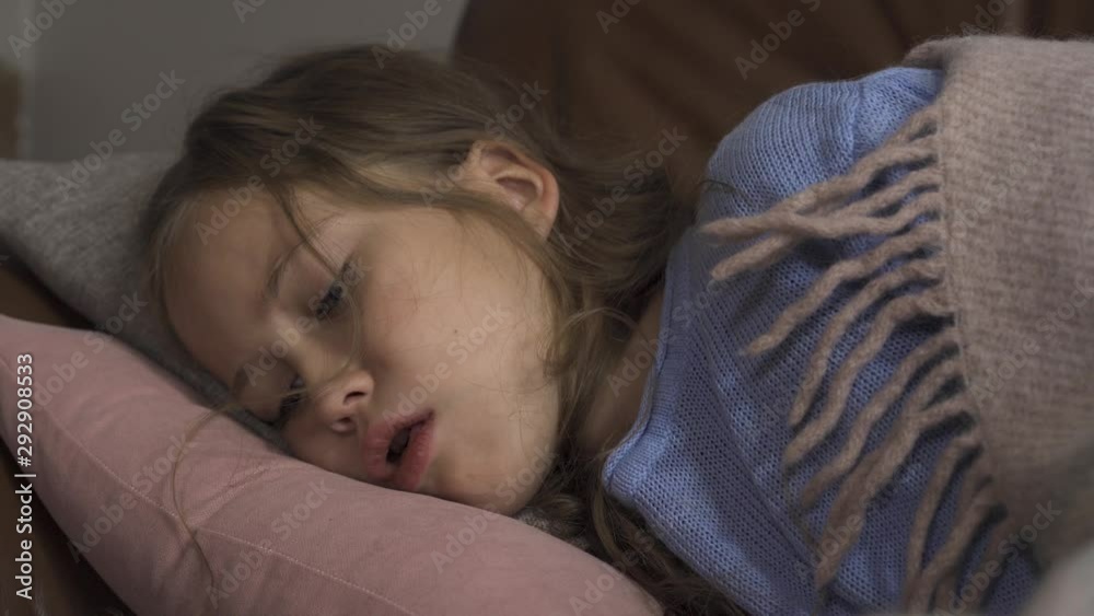 Sick young caucasian girl coughing while lying under blanket at home. The child has fever. Concept of health, illness, sickness, common cold, treatment Sick young caucasian girl coughing while lying under blanket at home. The child has fever. Concept of health, illness, sickness, common cold, treatment