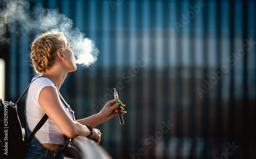 Young woman outdoor vaping e-cigarette on modern city buildings background photo
