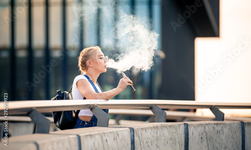 Young woman outdoor vaping e-cigarette on modern city buildings background photo