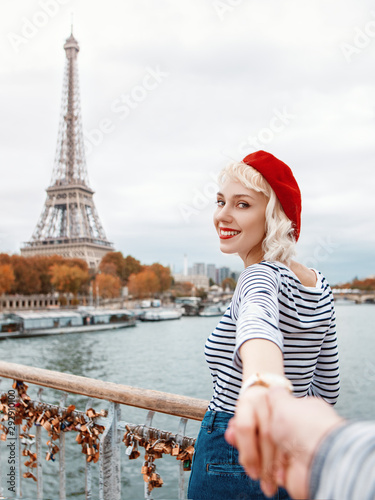 Follow me to Paris. Travel, tourism, advertising concept. Beautiful happy smiling girl wearing red beret, striped t-shirt holds her boyfriend by hand, walks on bridge with view of Eiffel tower. Autumn © Victoria Fox