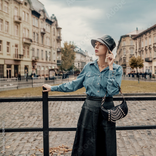 Outdoor autumn fashion portrait of elegant, luxury lady wearing trendy faux leather bucket hat, black midi skirt, denim shirt, holding small baguette bag with chains, posing in street of European city photo