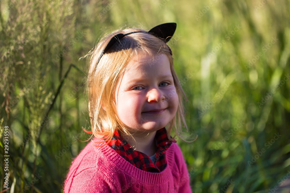 Horizontal closeup of smiling side lit pretty blond four-year old in pink sweater and cat ears headband enjoying a beautiful sunny fall golden hour morning outdoors Horizontal closeup of smiling side lit pretty blond four-year old in pink sweater and cat ears headband enjoying a beautiful sunny fall golden hour morning outdoors