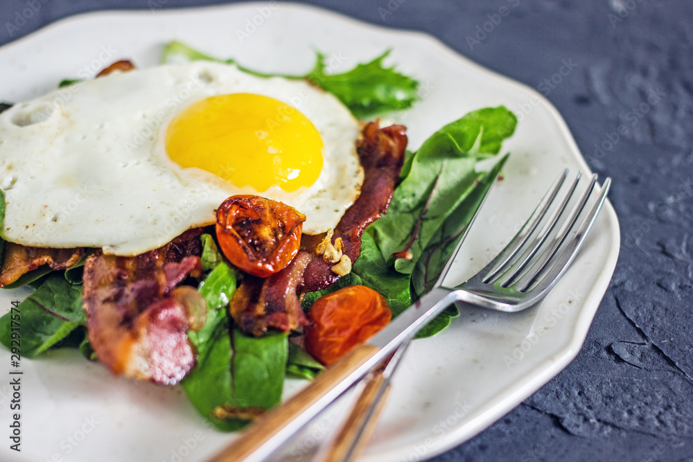 Appetizing breakfast with fried eggs and salad leaves. From above delicious meal with bacon egg tomatoes and green leaves served on white plate over gray textured background