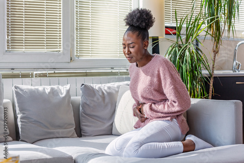 African American Woman in painful expression holding hands against belly suffering menstrual period pain, lying sad on home bed, having tummy cramp in female health concept photo