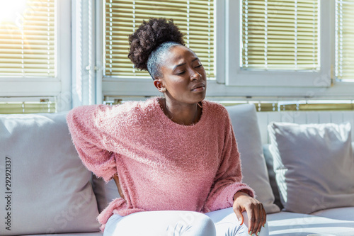 Young Black woman suffering from backache at home. Portrait of a young brunette girl sitting on the couch at home with a headache and back pain. Beautiful woman Having Spinal Or Kidney Pain photo