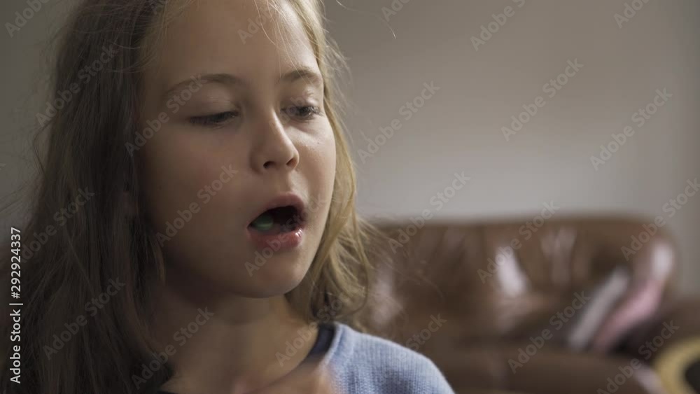 Close-up face of sick caucasian girl swallowing a green herbal pill and smiling while sitting at home. The child has fever. Concept of health, illness, sickness, common cold, treatment Close-up face of sick caucasian girl swallowing a green herbal pill and smiling while sitting at home. The child has fever. Concept of health, illness, sickness, common cold, treatment