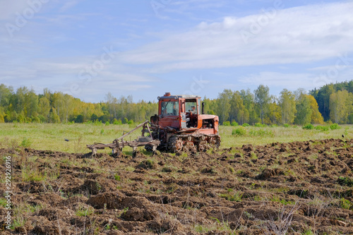 A tracked tractor with a plough plows the ground in autumn against the background of the forest.
