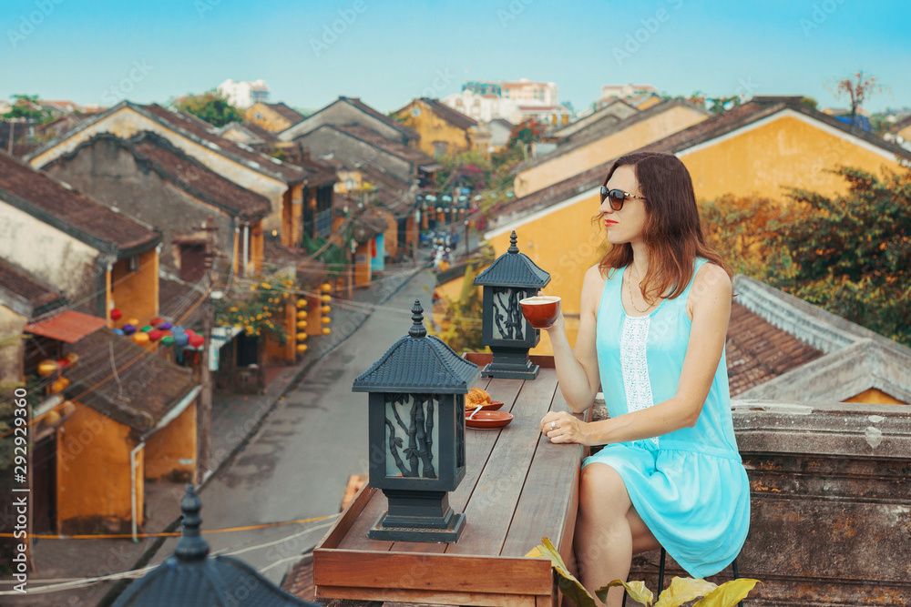 Tourist girl drinks coffee overlooking the old city of Hoi An, Vietnam. A young woman is enjoying the top view of the old Vietnamese city from the roof. A popular tourist destination in Vietnam