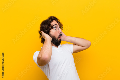 young crazy man with open mouth, looking horrified and shocked because of a terrible mistake, raising hands to head against yellow wall