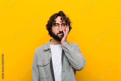 young crazy man feeling bored, frustrated and sleepy after a tiresome, dull and tedious task, holding face with hand against yellow wall