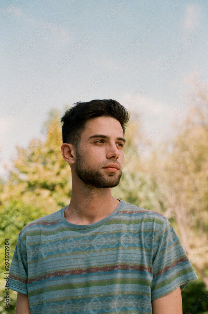 A handsome young brunette guy in a striped T-shirt stands against a background of plants and a blue sky and mysteriously looks into the distance