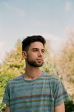 A handsome young brunette guy in a striped T-shirt stands against a background of plants and a blue sky and mysteriously looks into the distance