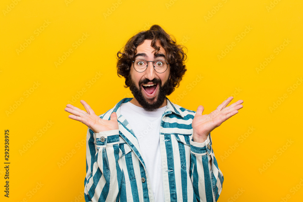 young crazy man looking happy and excited, shocked with an unexpected surprise with both hands open next to face against yellow wall - obrazy, fototapety, plakaty 