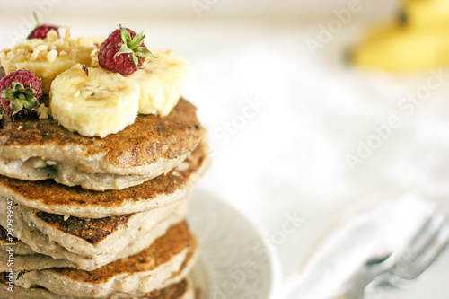 Delicious banana pancakes on white plate Stack of homemade appetizing fritters with sliced banana and fresh raspberries served on blurred white background