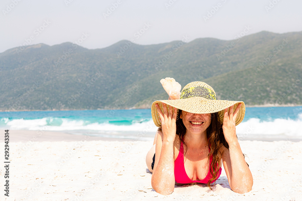 Woman in pink bikini and sun hat lying on the beach. Holding hat and laughing.