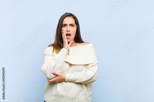 young pretty woman open-mouthed in shock and disbelief, with hand on cheek and arm crossed, feeling stupefied and amazed against blue background photo