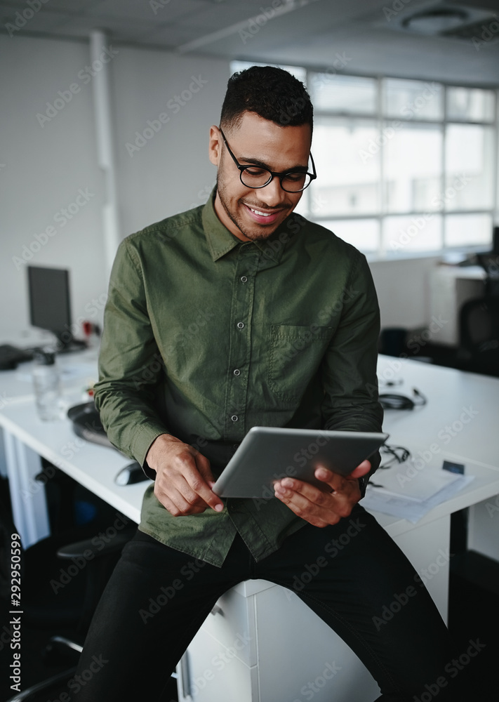 © StratfordProductions - Young businessman wearing eyeglasses sitting on the edge of office desk looking at digital tablet