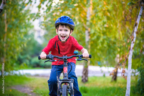 A small white Toddler boy in a protective helmet on his head sits on a children's bicycle. Toddler on a two-wheeled red bicycle looks forward. A sly smile on the child's face