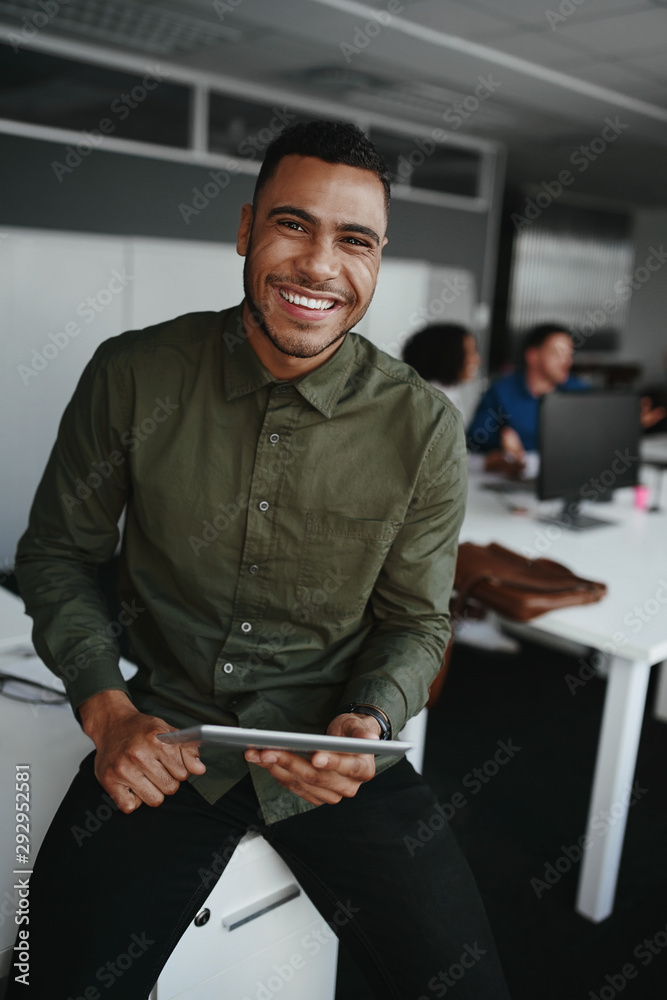© StratfordProductions - Portrait of a smiling young businessman holding digital tablet in hand looking to camera and colleague at background