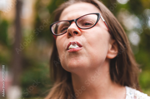 Young woman with eyeglasses chewing gum outdoors - Casually dressed girl with brown hair making bubble whit white gum in nature