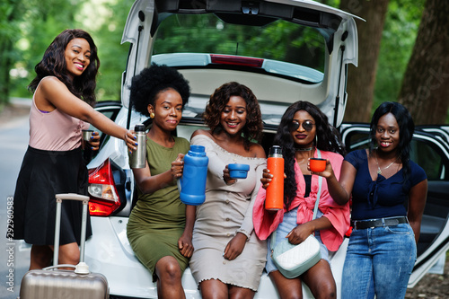 Group of five happy african american traveler girls sitting in car open trunk and drink tea from thermos.