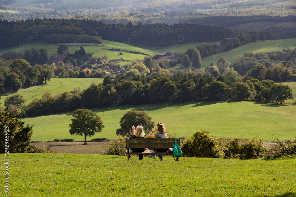Two women friends sitting and chatting on a bench in the afternoon sun overlooking the beautiful countryside of the Surrey hills from Newlands Corner