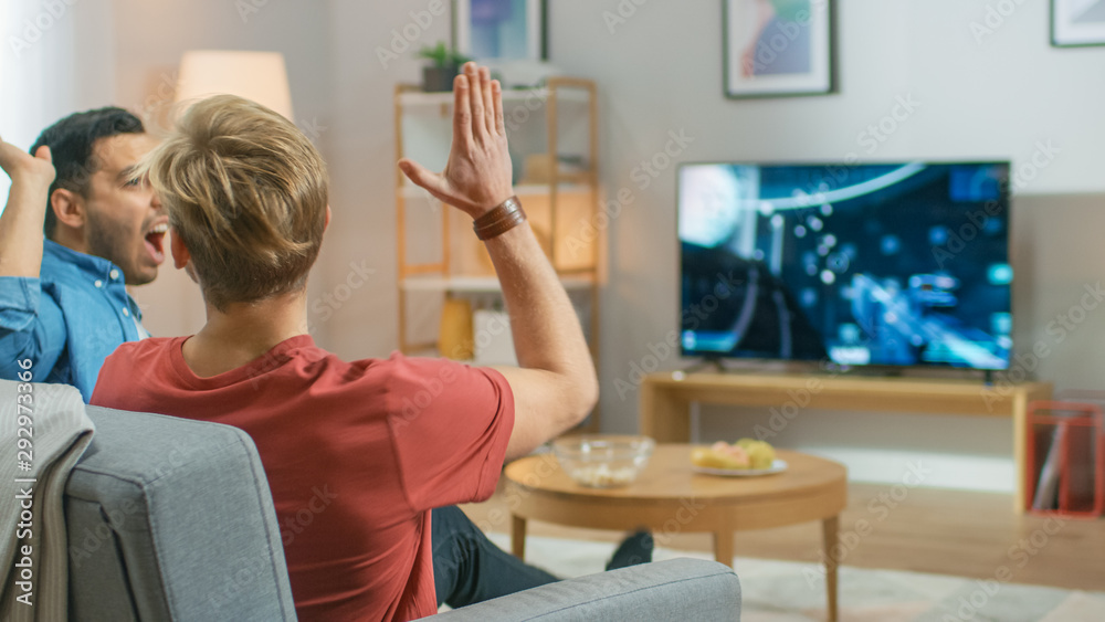 In the Living Room Two Friends Sitting on a Couch Holding Controllers and Doing High Five After Finishing Level in Competitive Video Game, 3D Action Shooter Gameplay Shown on TV Screen.