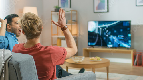 Картината върху платно In the Living Room Two Friends Sitting on a Couch Holding Controllers and Doing High Five After Finishing Level in Competitive Video Game, 3D Action Shooter Gameplay Shown on TV Screen