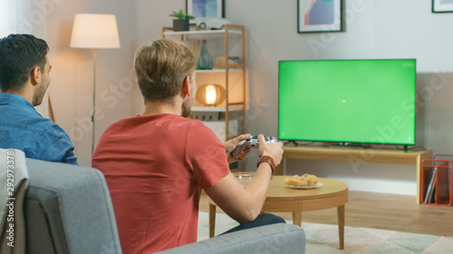 In the Living Room Two Friends Sitting on a Couch Holding Controllers Playing Competitive Video Game on a Green Chroma Key TV Screen. 