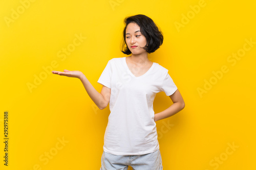 Asian young woman over isolated yellow wall holding copyspace with doubts