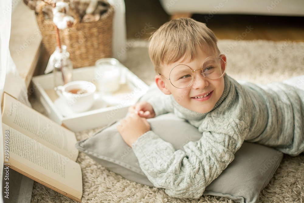 A beautiful little boy in a knitted sweater is reading by the window with a mug of hot tea. Cozy. Autumn. A beautiful little boy in a knitted sweater is reading by the window with a mug of hot tea. Cozy. Autumn.