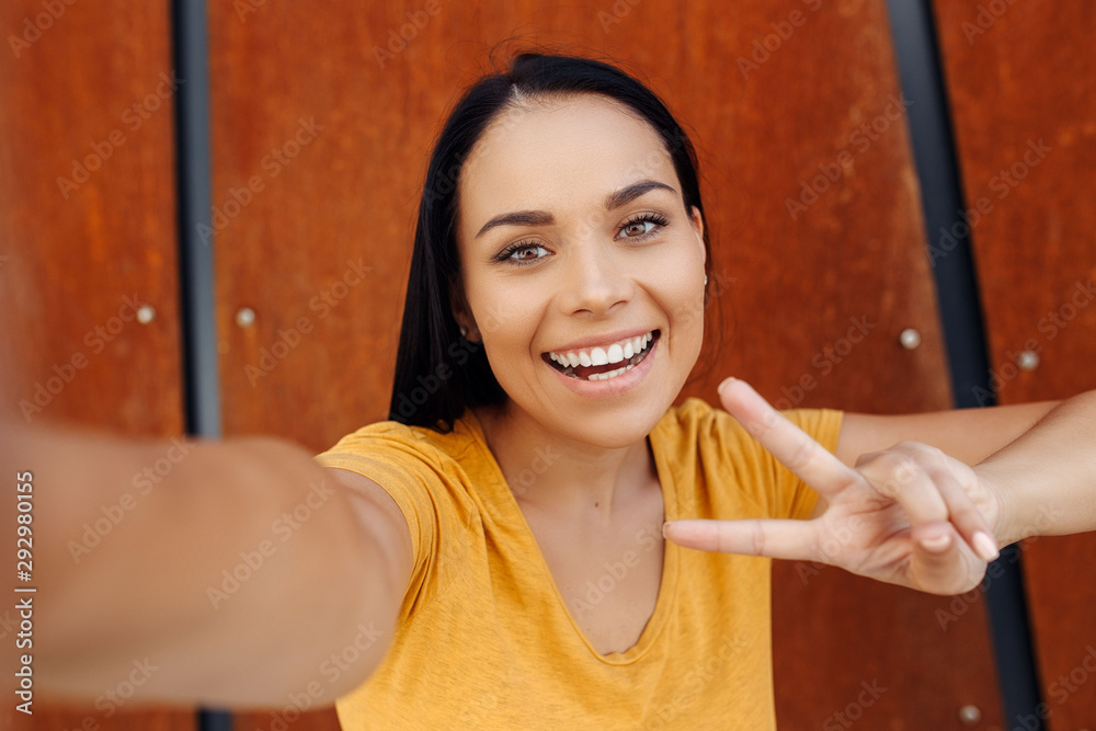 Attractive brunette young woman with bright smile dressed in casual clothes doing selfie on smartphone and show peace gesture in front of rusty wall. Attractive brunette young woman with bright smile dressed in casual clothes doing selfie on smartphone and show peace gesture in front of rusty wall.