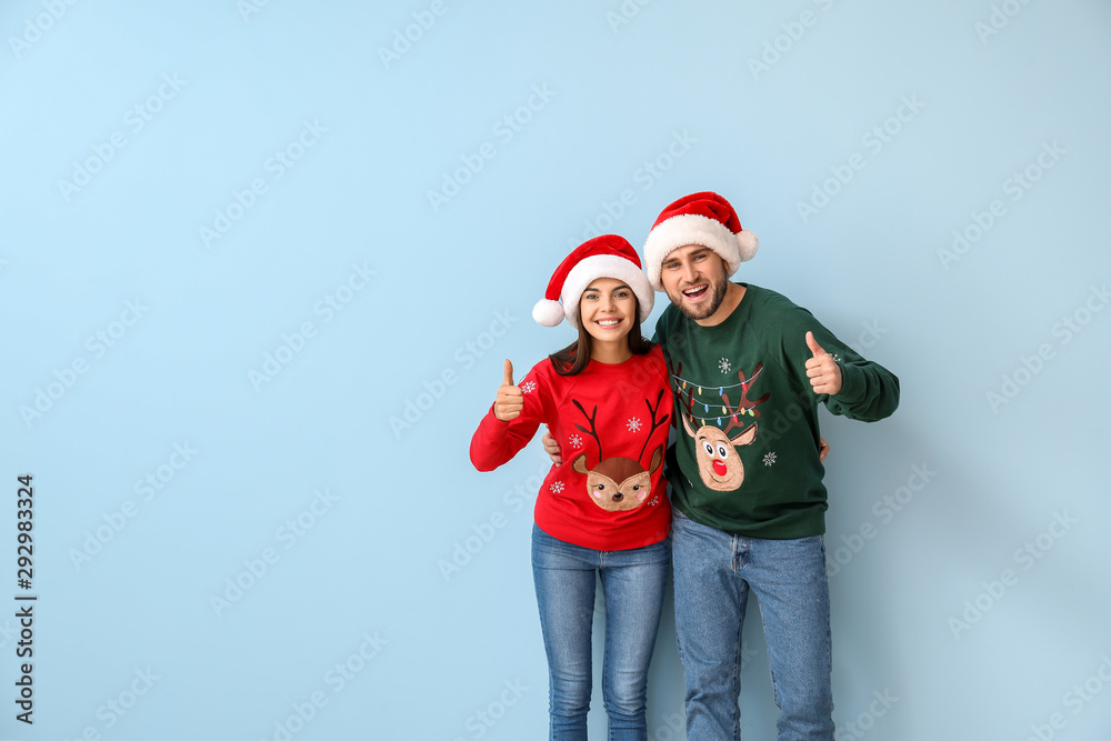 Happy couple in Christmas sweaters and Santa hats showing thumb-up on color background