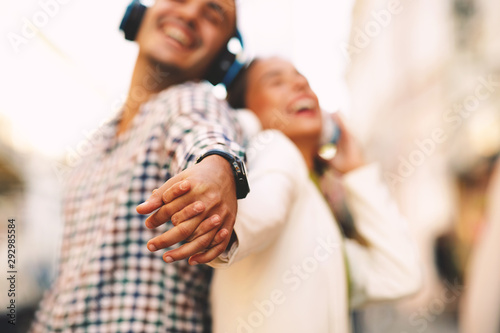 Happy young couple dancing on the street and listening to music through headphones and holding hands. Selective focus on hadns.