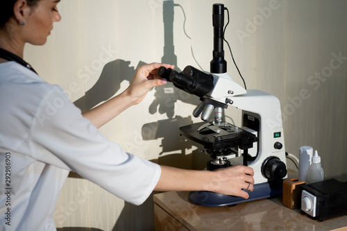 female doctor in white medical uniform with stethoscope on neck adjusts the microscope in laboratory photo