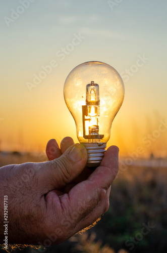  Man holding halogen light bulb in front of the sun , at golden hour to make it appear bright. Concept for green energy , solar energy , environment friendly.