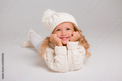 Winter clothes. Portrait of a little curly-haired girl in a knitted white winter hat. little blonde girl in white knitted hat and sweater smiling light background isolate, space for text