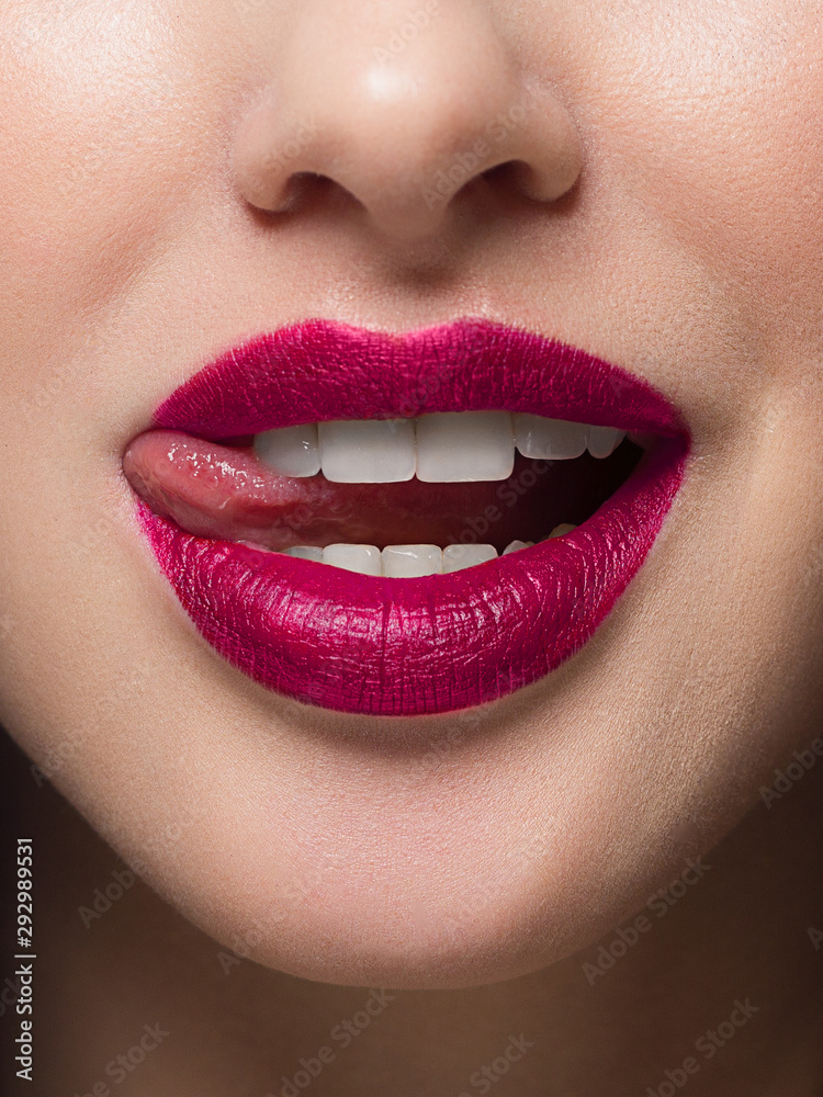 Fototapeta premium Close-up of sexy female lips with tongue. Clean skin and a clear lip contour are outlined with a fashionable marsala lipstick. White teeth and the beauty of smile for stamotologii, spa or cosmetology