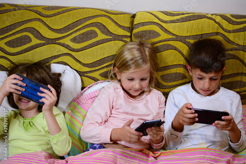 Top View of Adorable children playing with cell phones in bed covered with blankets. Kids enjoy playing games via smartphone. Kids love to play with phones in the morning.