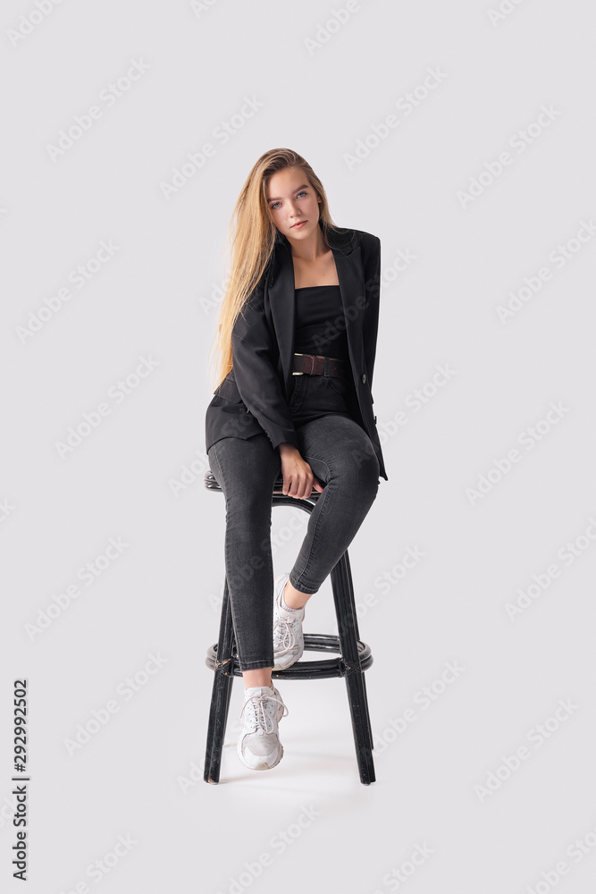 Fototapeta premium Full length portrait of beautiful young girl sitting on tall chair in black jeans and jacket over gray background